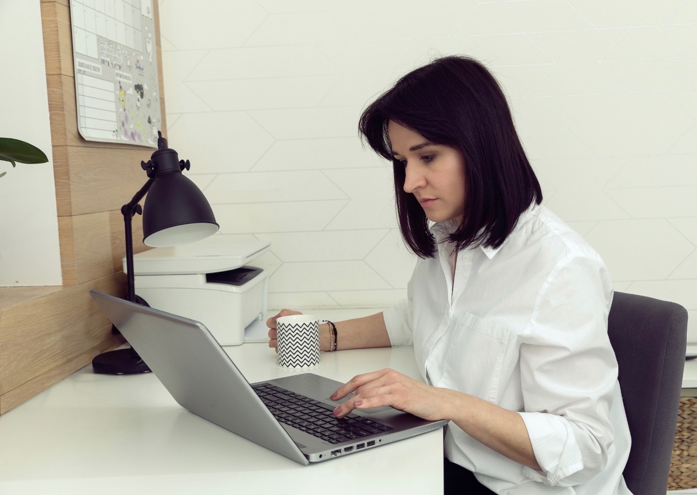 Kobieta w białej koszuli skupiona na pracy przy laptopie, z kubkiem w zygzaki obok, biurko z lampką, drukarką i kalendarzem w tle.