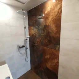 Nowoczesna kabina prysznicowa z deszczownicą, wykończona płytkami imitującymi rdzę na jednej ścianie oraz jasnymi płytkami na pozostałych ścianach, z widocznym odpływem liniowym w podłodze...