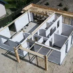 Wznoszone ściany parteru domu jednorodzinnego z bloczków betonu komórkowego, widok z drona, szalunki pod wieniec, częściowo zadaszona konstrukcja tarasu.