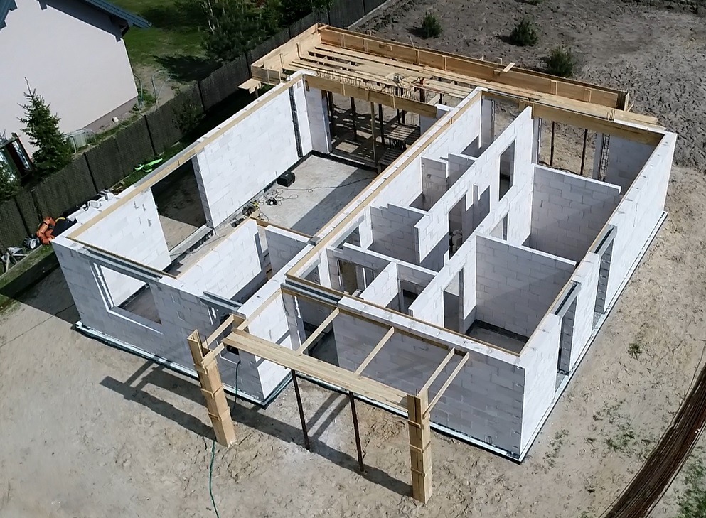 Wznoszone ściany parteru domu jednorodzinnego z bloczków betonu komórkowego, widok z drona, szalunki pod wieniec, częściowo zadaszona konstrukcja tarasu.