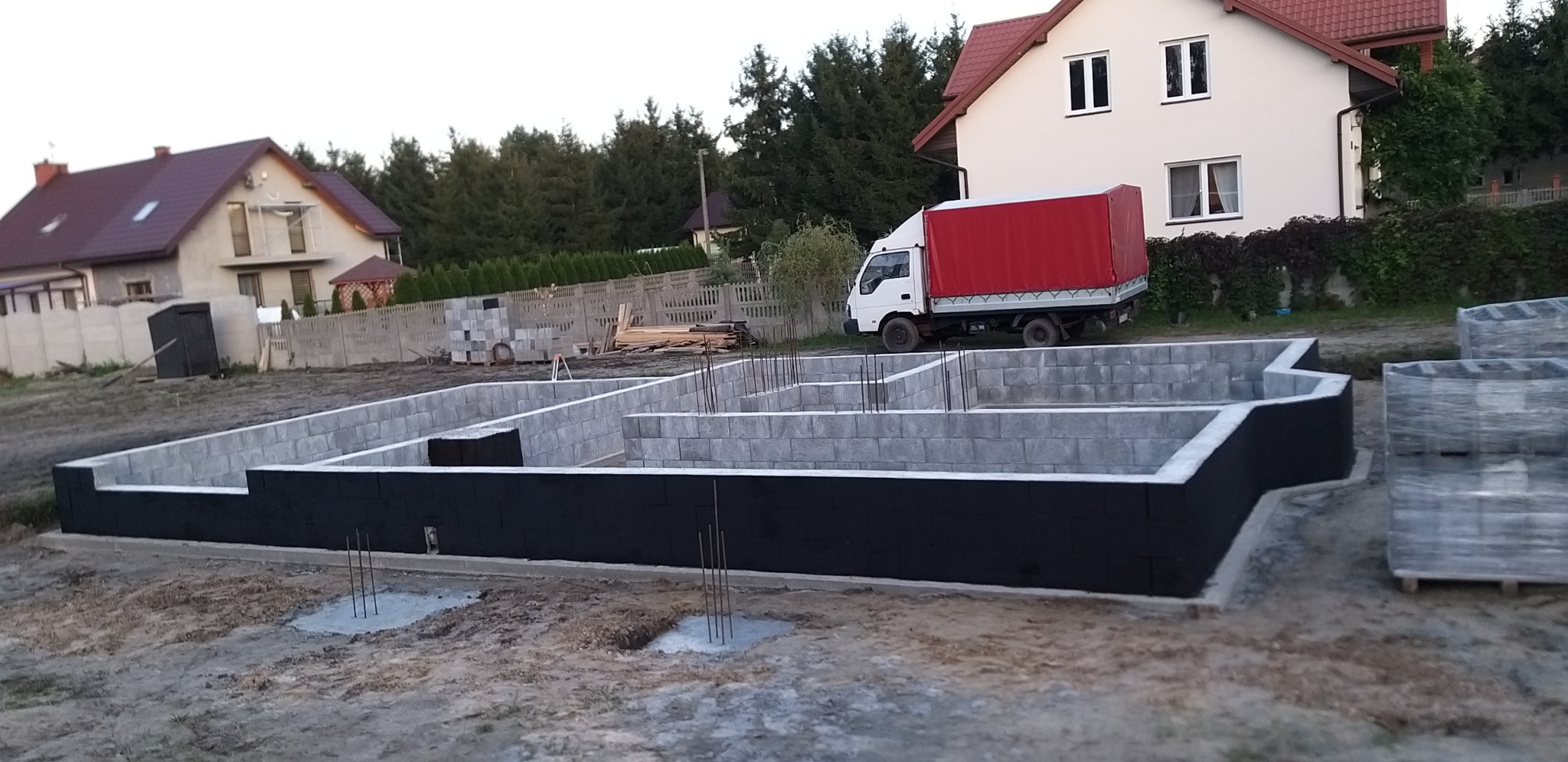 Zalane fundamenty z wystającymi prętami zbrojeniowymi, ściany fundamentowe z bloczków betonowych zabezpieczone czarną izolacją, w tle dom w trakcie budowy i samochód dostawczy z czerwoną plandeką.