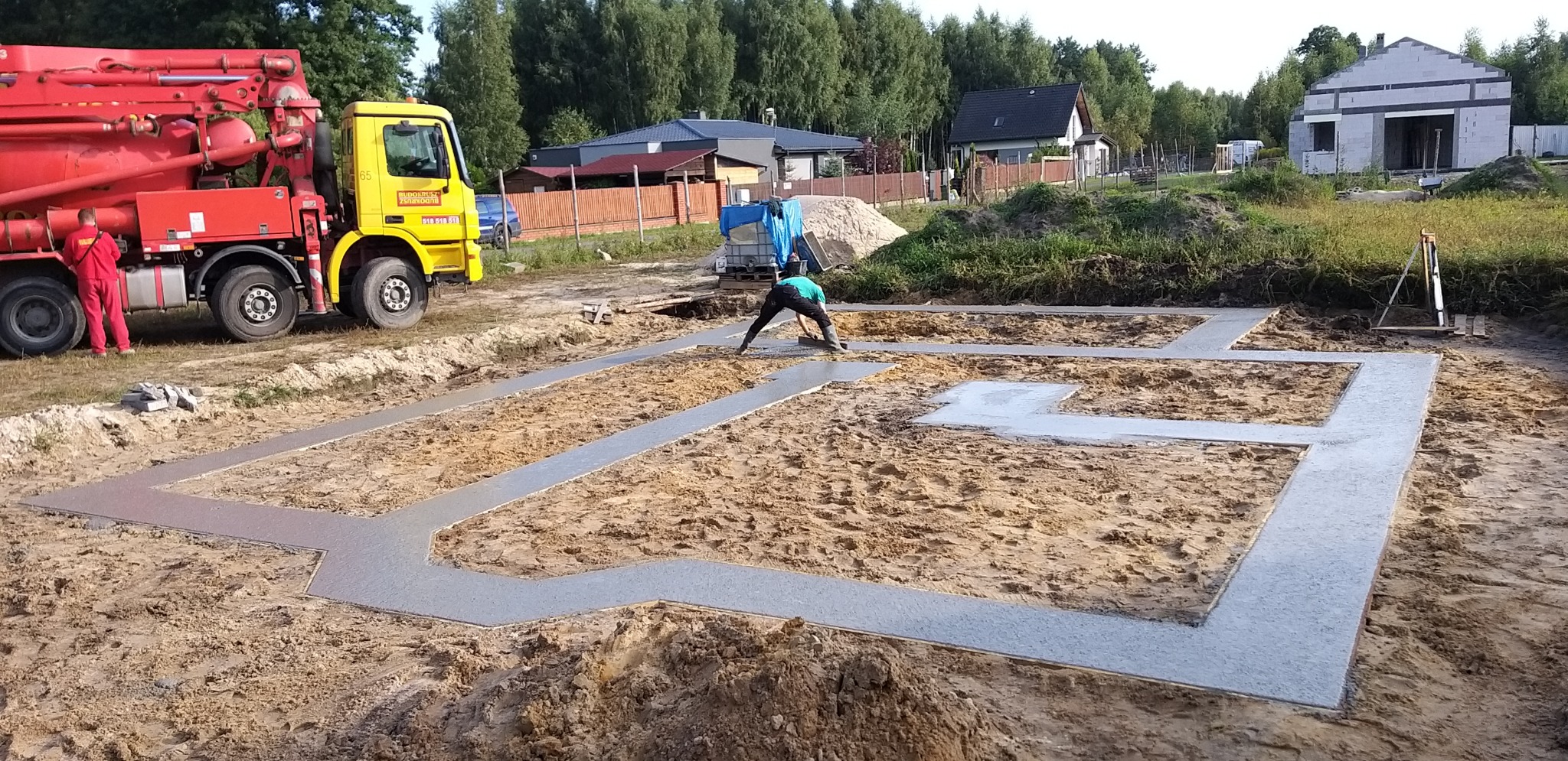 Wylewanie betonowych fundamentów pod dom jednorodzinny; widoczny pracownik wyrównujący beton, żółto-czerwona betoniarka z tyłu, w tle dom w trakcie budowy z białego bloczka.