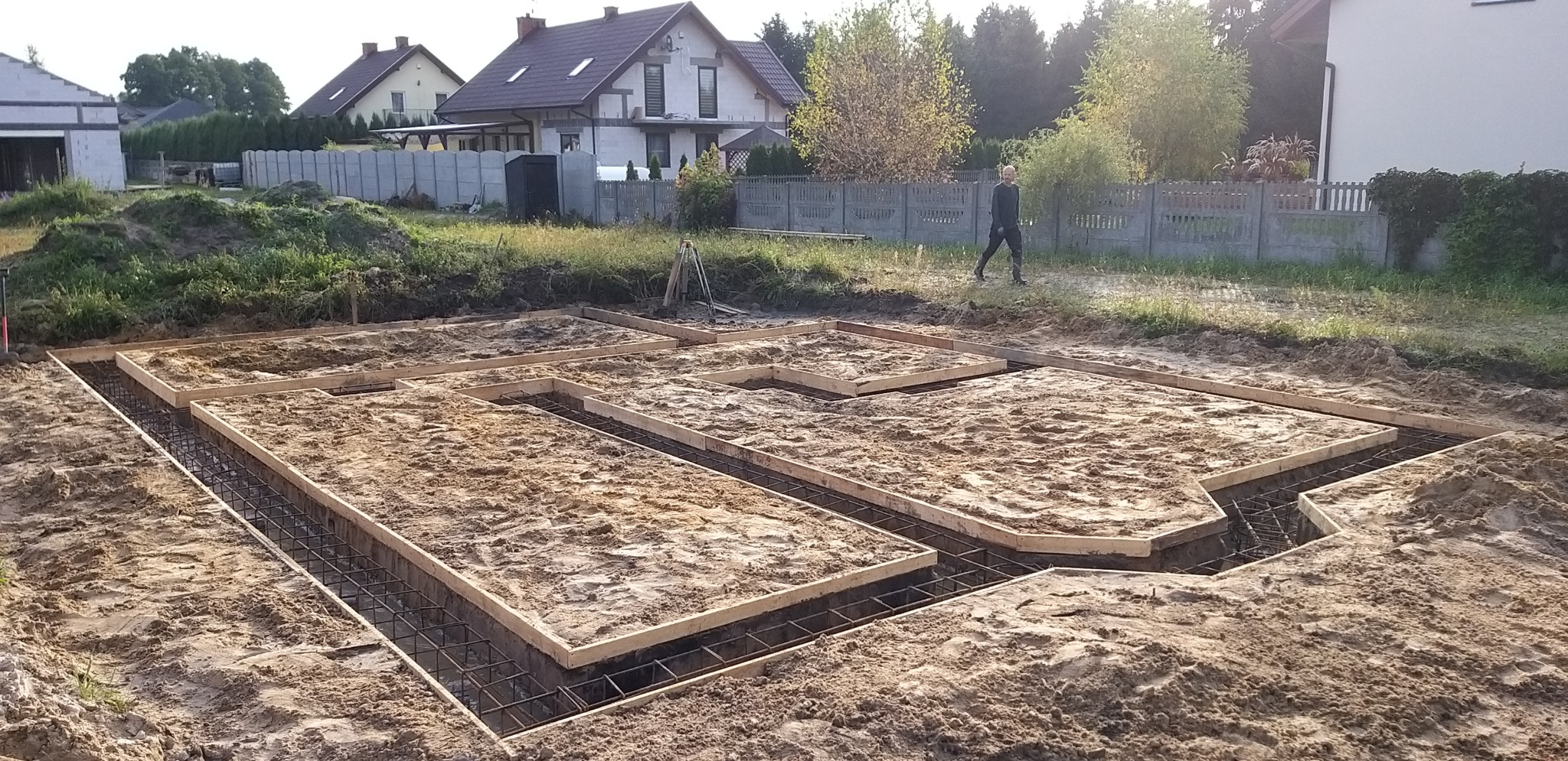 Zalane piaskiem szalunki zbrojeniowe pod fundamenty domu jednorodzinnego na działce z widocznym zarysem budynku w tle, osoba w oddali.