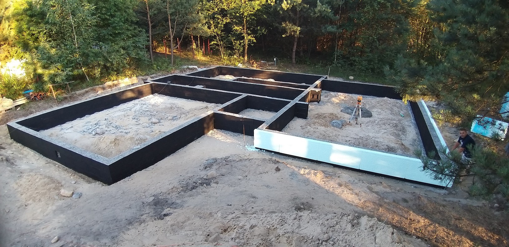 Fundament domu w trakcie budowy, widoczne czarne ściany fundamentowe, białe płyty izolacyjne, piasek wewnątrz, niwelator laserowy na trójnogu i pracownik siedzący obok.