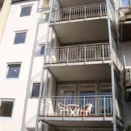 Wielopiętrowy budynek z balkonami z metalowymi barierkami, widok z dołu na bocznej elewacji, na jednym z balkonów białe krzesła i stolik.