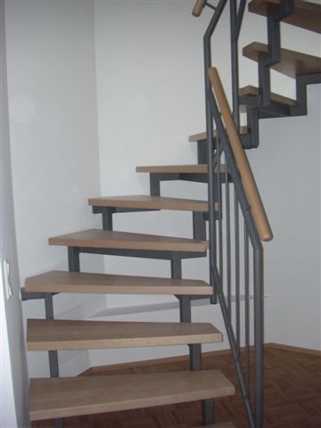 Schody wewnętrzne z drewnianymi stopniami i metalową konstrukcją nośną oraz poręczą z elementami drewnianymi, zamontowane w jasnym pomieszczeniu.