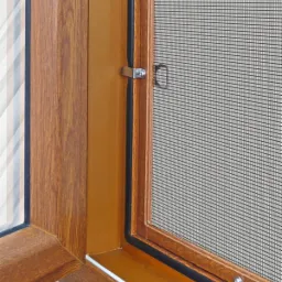 Fragment okna z drewnopodobną ramą i zamontowaną moskitierą z uchwytem zabezpieczającym, widoczne uszczelki.