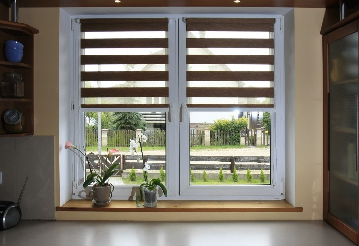 Okno z dwoma skrzydłami, przesłonięte roletami w brązowo-beżowe pasy, na parapecie kwiaty w doniczkach, za oknem widok na ogród i ulicę.