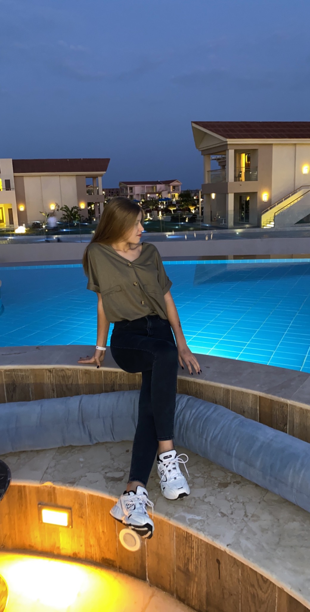 Młoda kobieta siedzi na krawędzi podświetlonego basenu w wieczornej scenerii, w tle budynki z oświetleniem.