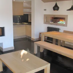 Jasne, minimalistyczne wnętrze z drewnianą podłogą, stołem, ławką i kominkiem w ścianie, widoczna kuchnia w tle.