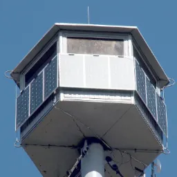 Wieża obserwacyjna z panelami fotowoltaicznymi zamontowanymi na ścianach, widok od dołu na tle błękitnego nieba.