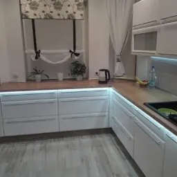 Nowoczesna kuchnia z białymi szafkami, drewnianym blatem i podświetleniem LED pod szafkami wiszącymi oraz nad blatem dolnych szafek, widoczne gniazdka elektryczne nad blatem.