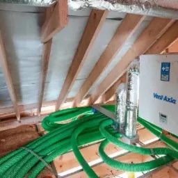 Centrala wentylacyjna Vent-Axia z podłączonymi przewodami wentylacyjnymi w kolorze zielonym, zamontowana na poddaszu z widoczną izolacją i drewnianą konstrukcją dachu.