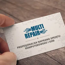 Wizytówka firmy Multi Repair, specjalizującej się w profesjonalnej naprawie sprzętu komputerowego i GSM, trzymana w palcach na tle drewnianego stołu.
