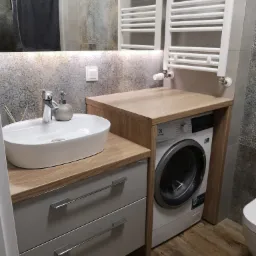 Nowoczesna łazienka z umywalką nablatową, szafką z szufladami, pralką zabudowaną pod blatem drewnianym i grzejnikiem drabinkowym.