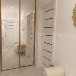 Korytarz z dużą szafą przesuwną z lustrzanymi drzwiami w romby, odbijającymi wnętrze pokoju, białe drzwi z poziomymi paskami i kremowa pufa z kryształkami.