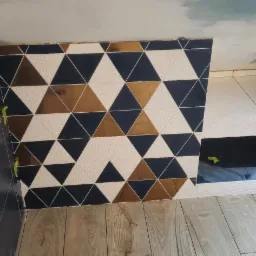 Fragment ściany wyłożonej trójkątnymi płytkami w kolorach białym, granatowym i miedzianym, z widocznymi żółtymi klinami poziomującymi oraz fragmentem drewnopodobnej podłogi.