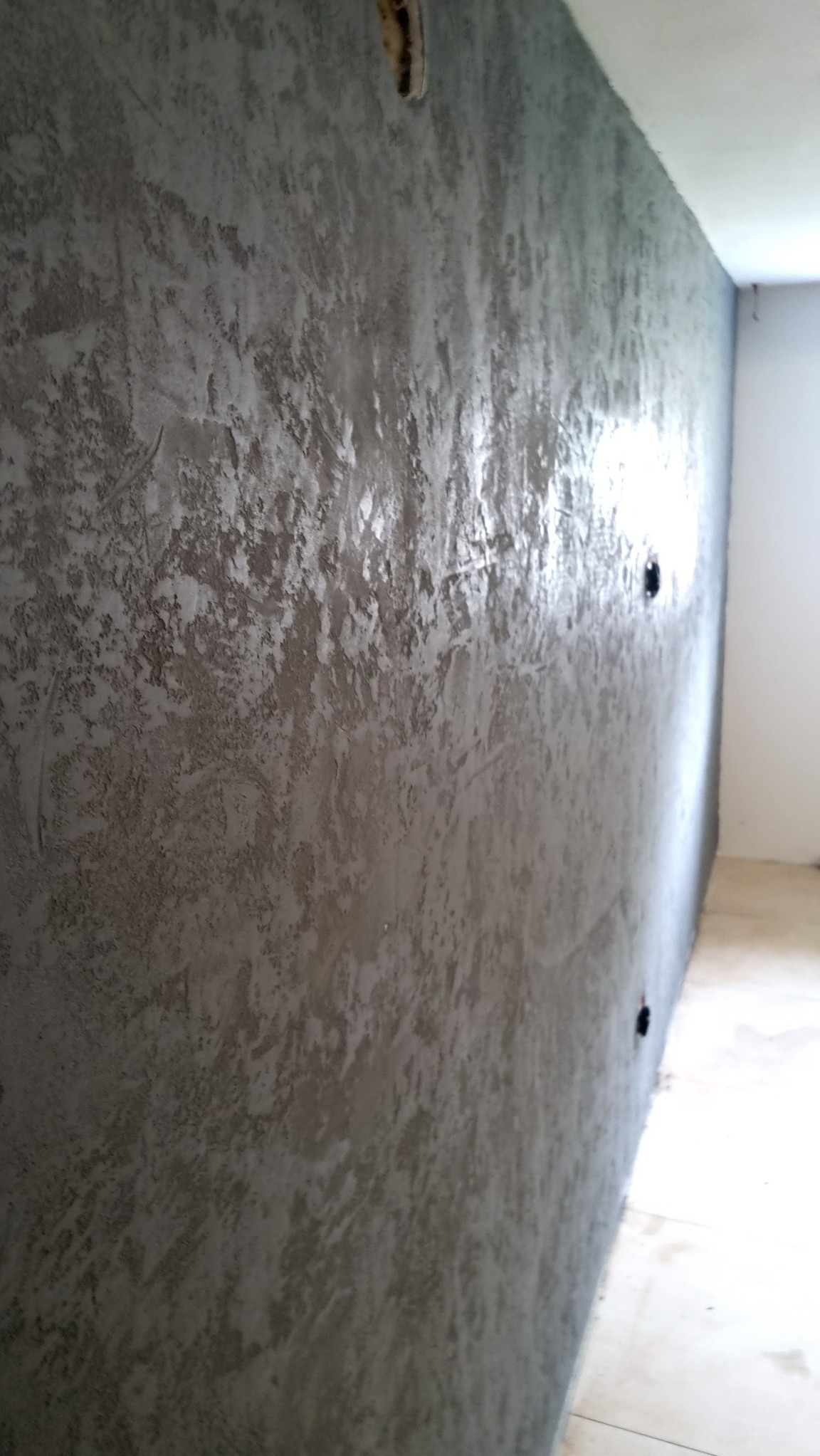 Ściana pokryta dekoracyjnym tynkiem w odcieniach szarości, z widocznymi fakturami i refleksami światła, z elementami instalacji elektrycznej w górnej części.