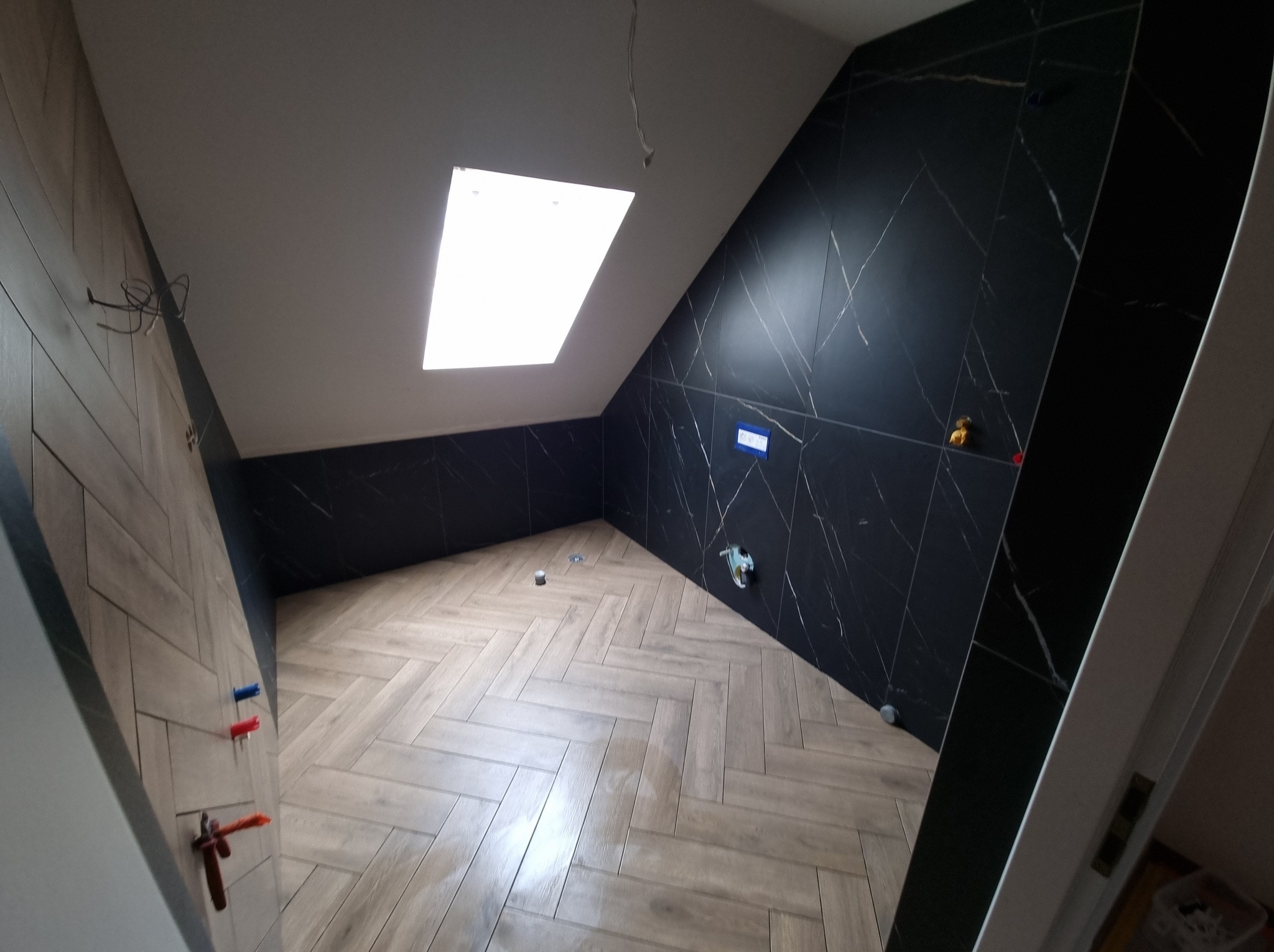 Wykończona łazienka na poddaszu z czarnymi płytkami imitującymi marmur na ścianach i jasnymi płytkami ułożonymi w jodełkę na podłodze, oświetlona przez okno dachowe.