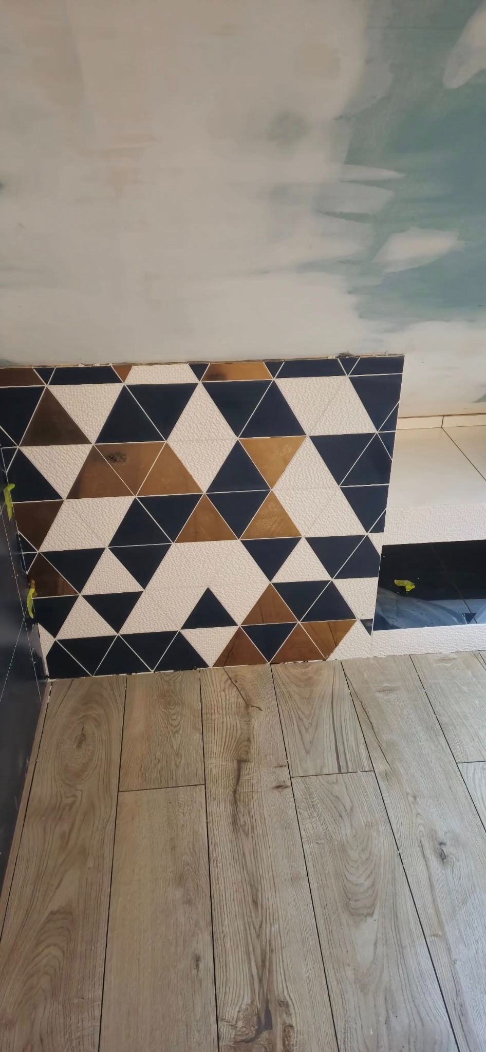 Fragment ściany wyłożonej trójkątnymi płytkami w kolorach białym, granatowym i miedzianym, z widocznymi żółtymi klinami poziomującymi oraz fragmentem drewnopodobnej podłogi.