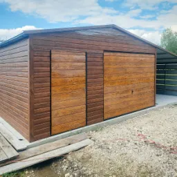 Brązowy garaż blaszany z imitacją drewna, z dwoma przesuwnymi drzwiami, małym oknem po lewej stronie i betonową wylewką, widoczny fragment zadaszenia z tyłu, na ziemi gruz i deski.