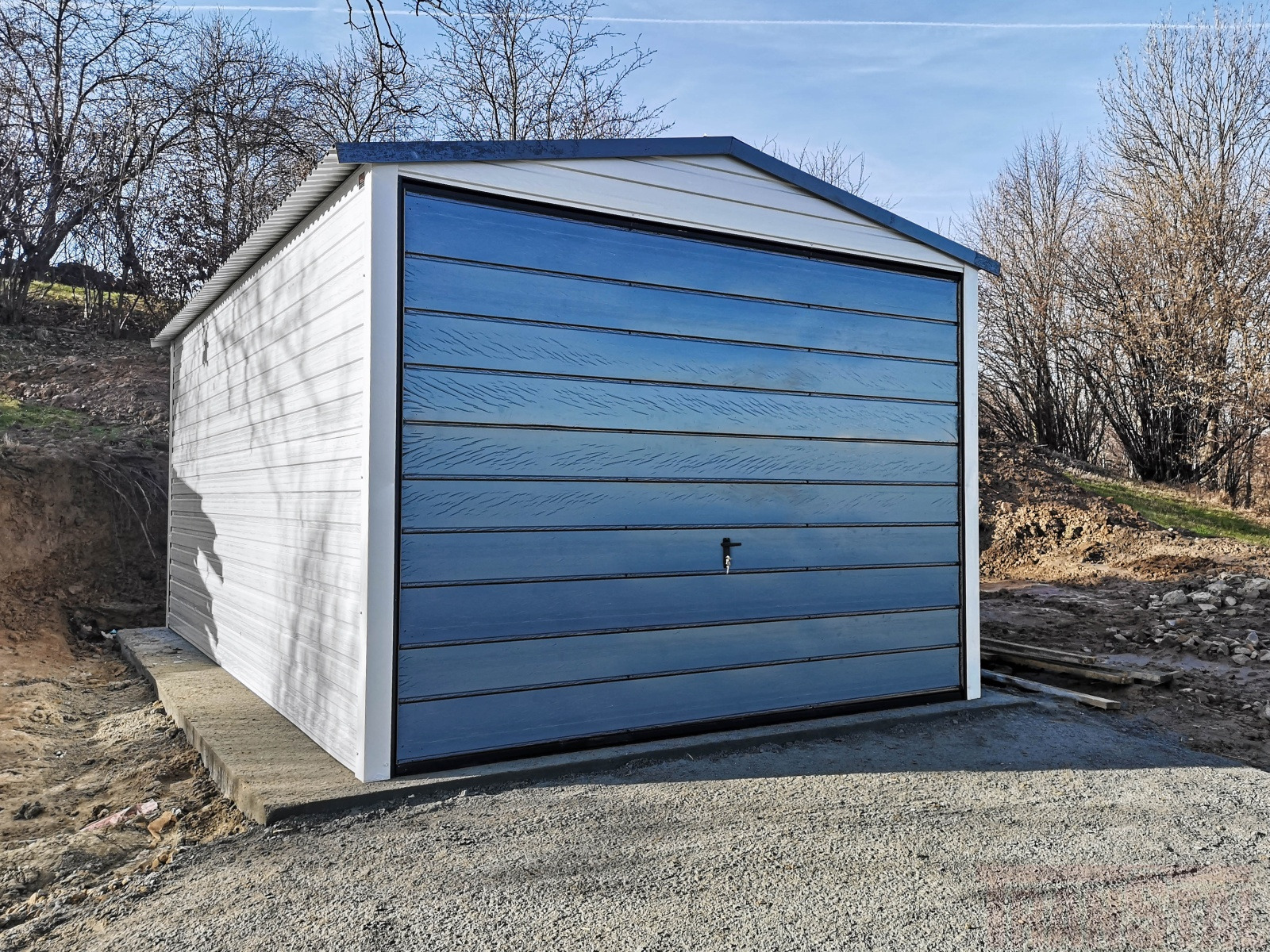 Gotowy garaż blaszany z bramą segmentową w kolorze grafitowym, umieszczony na betonowej wylewce, z widocznymi drzewami bezlistnymi w tle.