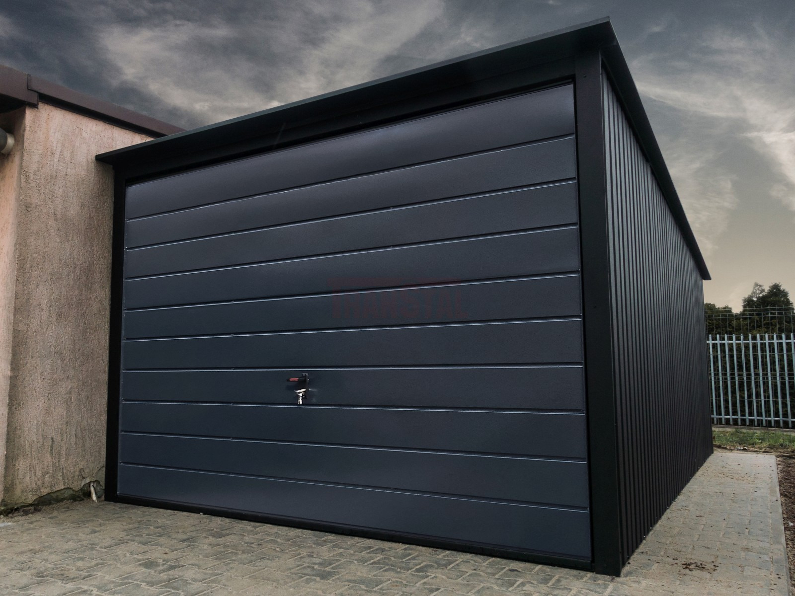 Nowoczesny, grafitowy garaż blaszany z poziomymi panelami, zamkiem na klucz w drzwiach, przylegający do budynku z jasną elewacją, widok z zewnątrz na tle pochmurnego nieba i ogrodzenia.