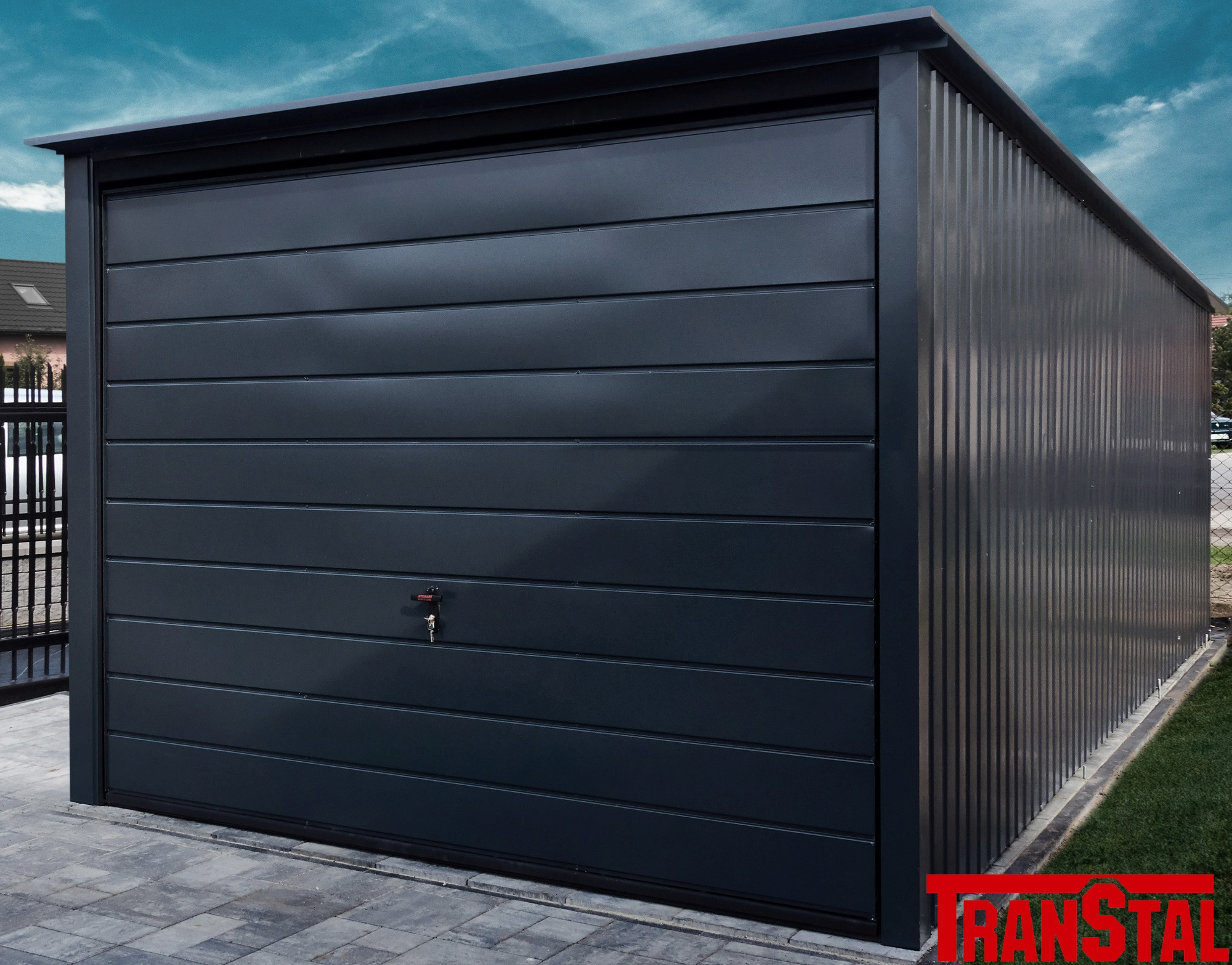 Nowoczesny, antracytowy garaż blaszany z poziomymi panelami na froncie i pionowymi żłobieniami po bokach, zamek z kluczem w drzwiach, kostka brukowa przed garażem, logo producenta w rogu.