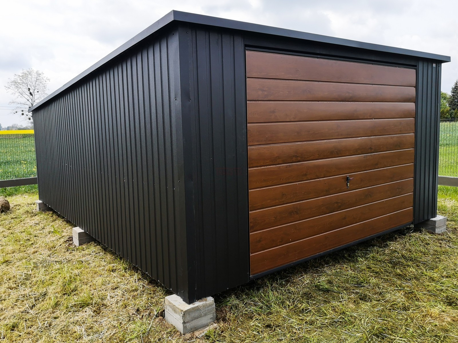 Gotowy garaż blaszany z brązowymi drzwiami segmentowymi, umieszczony na betonowych bloczkach na trawie, widok z boku.