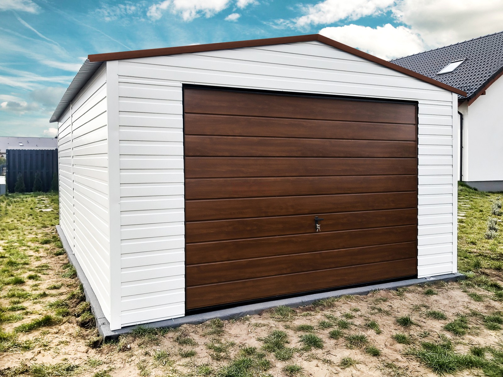 Gotowy garaż blaszany z brązową bramą segmentową i białymi ścianami, zamek w bramie, na trawiastym podłożu, w tle dom jednorodzinny.