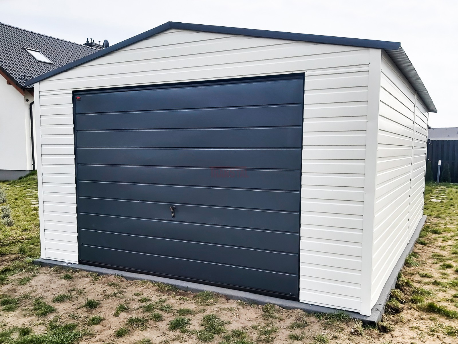 Gotowy garaż blaszany z bramą segmentową w kolorze grafitowym, zlokalizowany na działce z kostką obrzeżową.