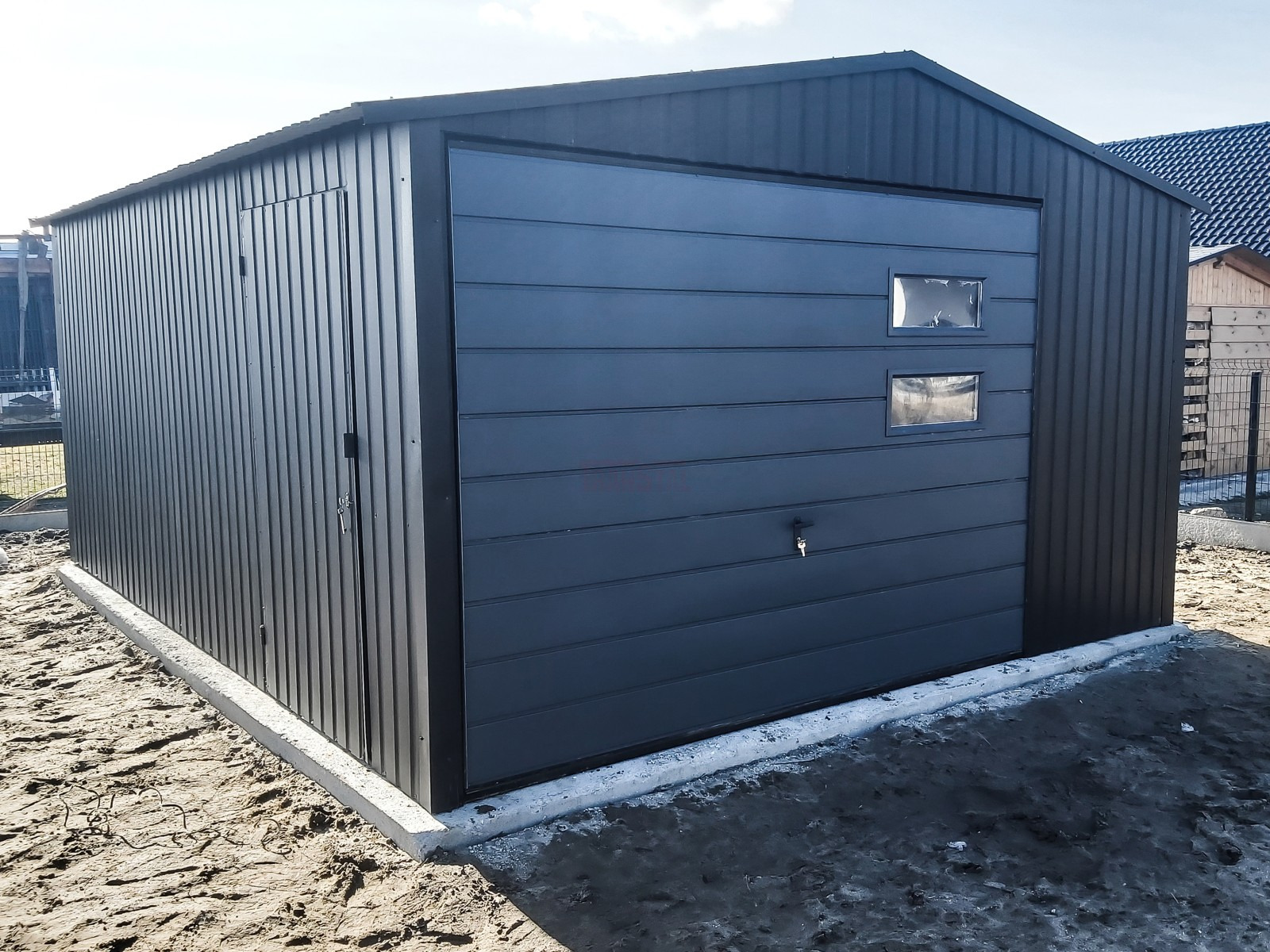Gotowy, ciemnoszary garaż blaszany z bramą segmentową z dwoma okienkami, osadzony na betonowej wylewce.