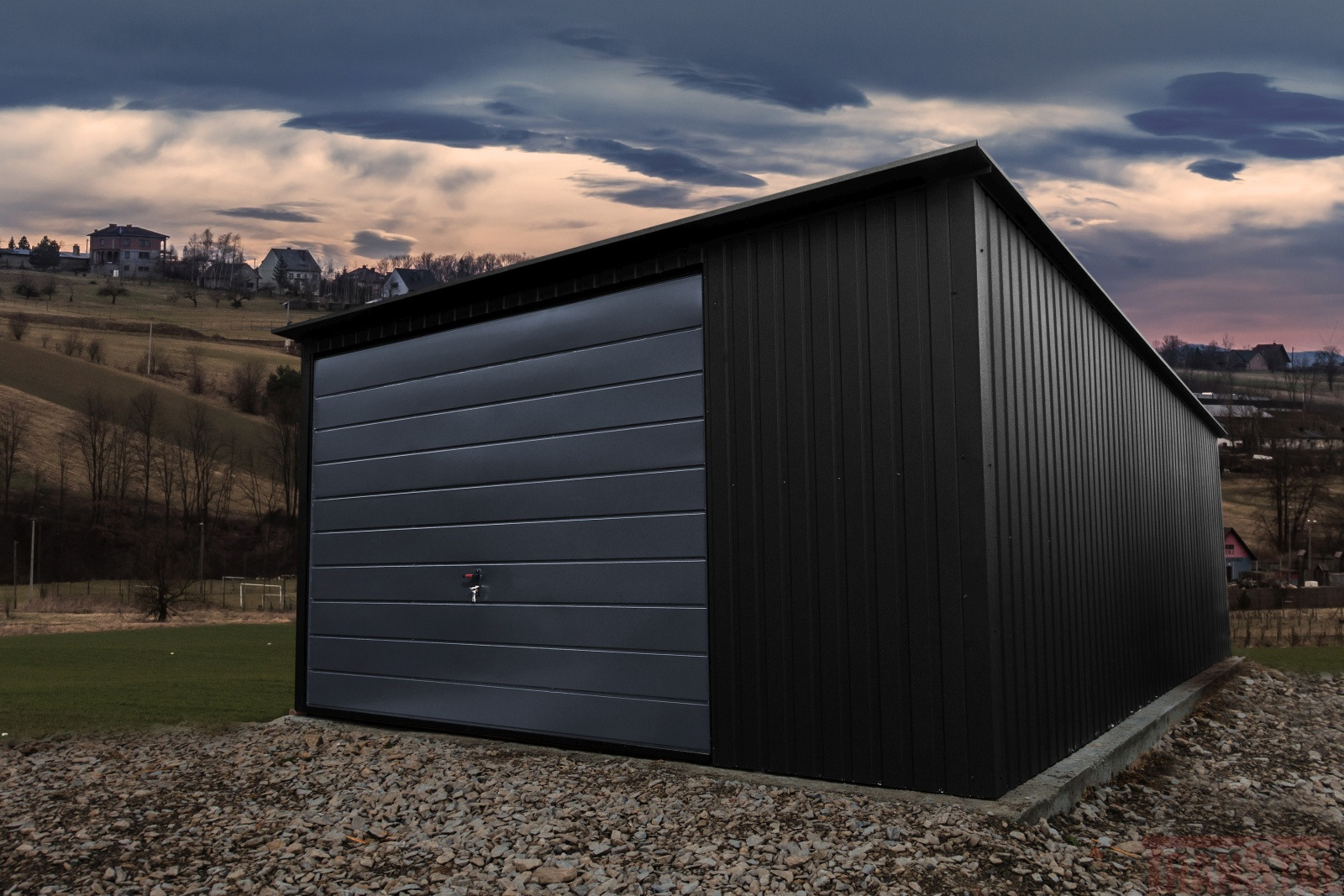 Nowoczesny, grafitowy garaż blaszany z bramą segmentową, usytuowany na żwirowym podłożu, z widokiem na pagórkowaty krajobraz i zachmurzone niebo o zachodzie słońca.