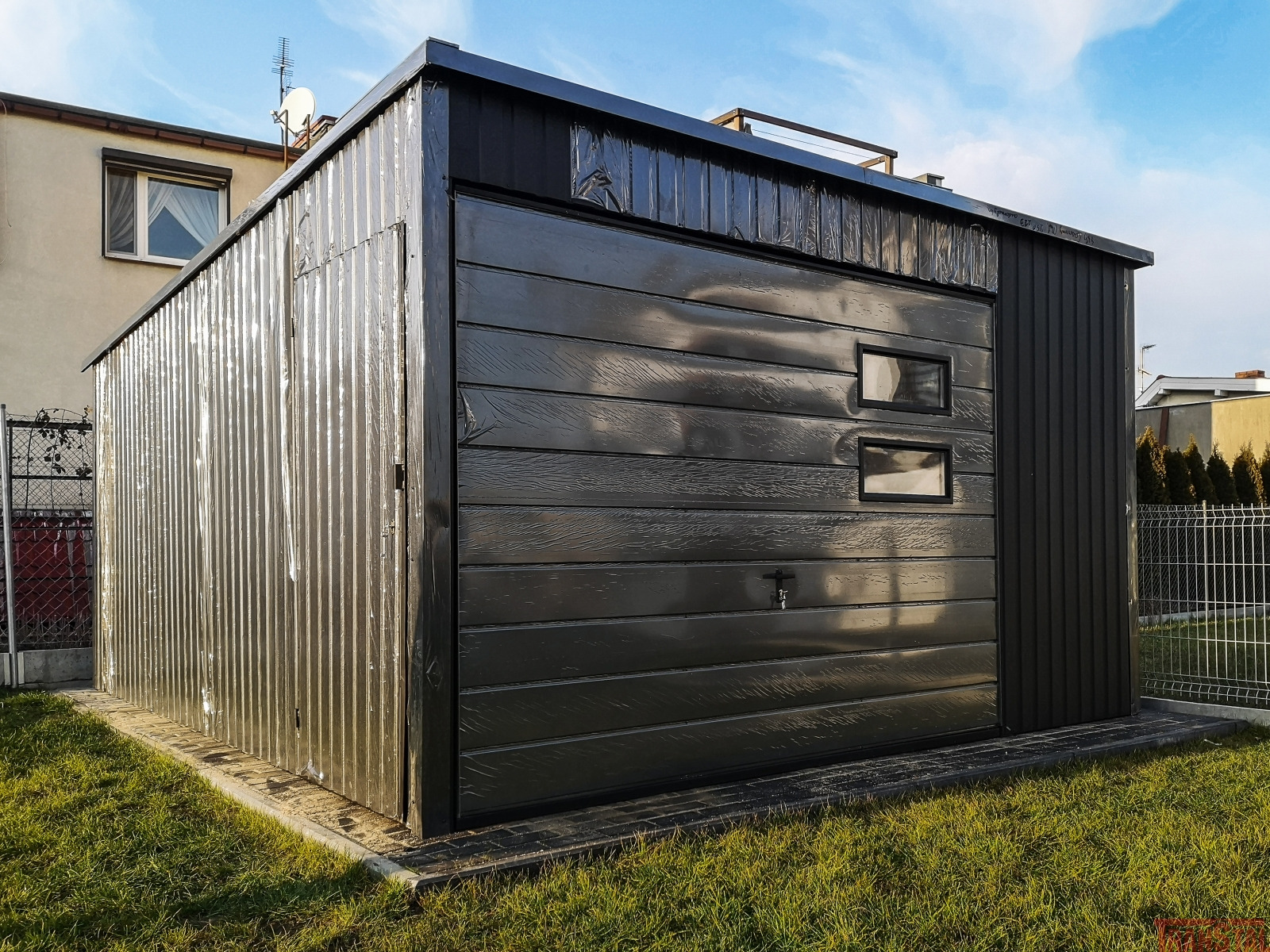 Nowoczesny, czarny garaż z blachy trapezowej i segmentową bramą z dwoma małymi okienkami, na trawniku przed domem.
