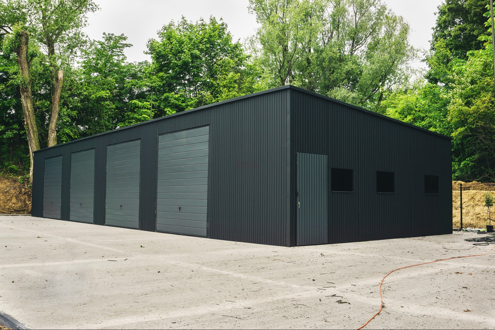 Czarny budynek z blachy falistej z czterema szarymi bramami garażowymi i drzwiami wejściowymi, otoczony zielenią drzew