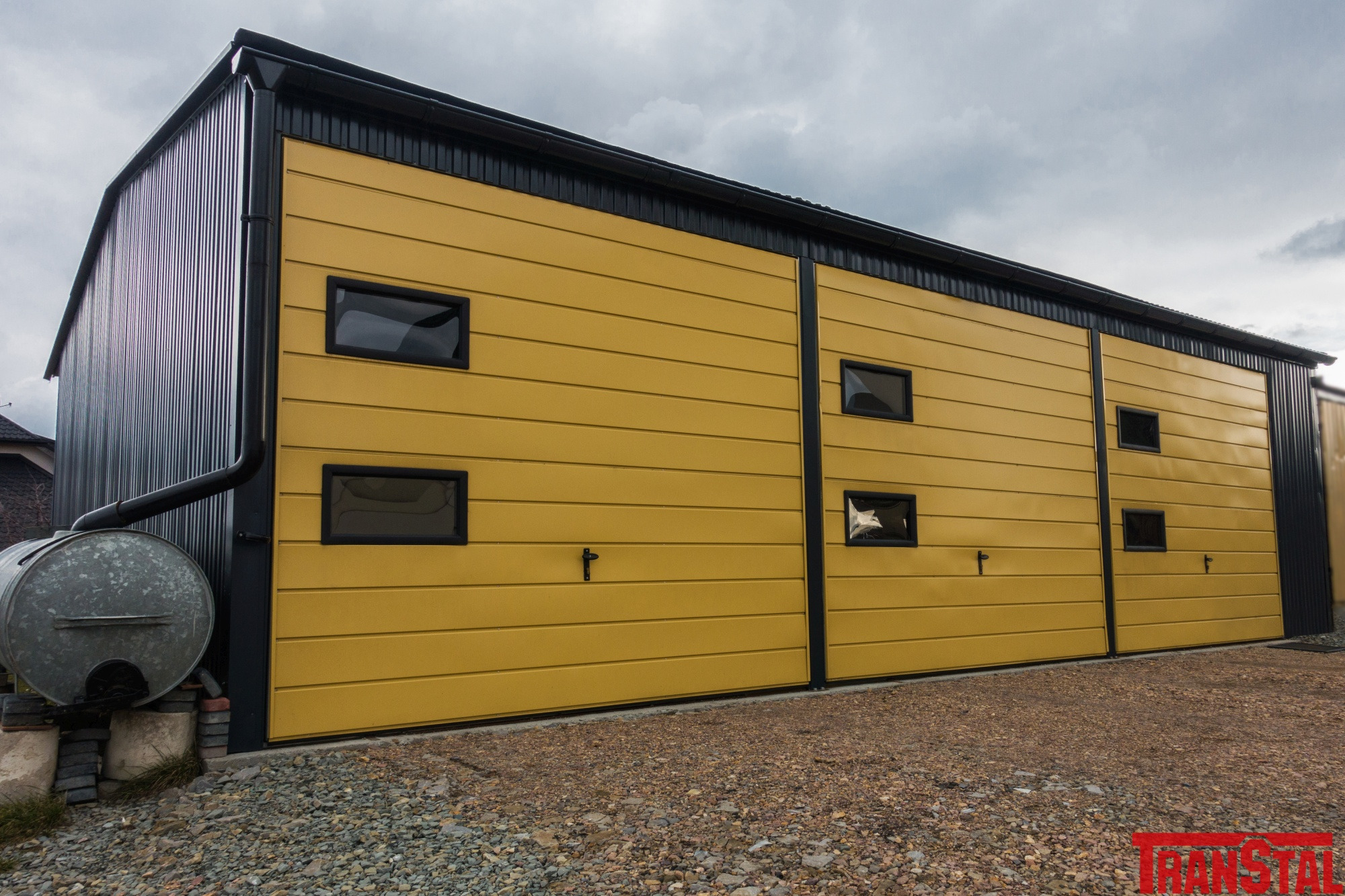 Żółto-czarna hala z blachy trapezowej z czterema małymi oknami i zbiornikiem na deszczówkę z boku, widok z zewnątrz na żwirowym podłożu.