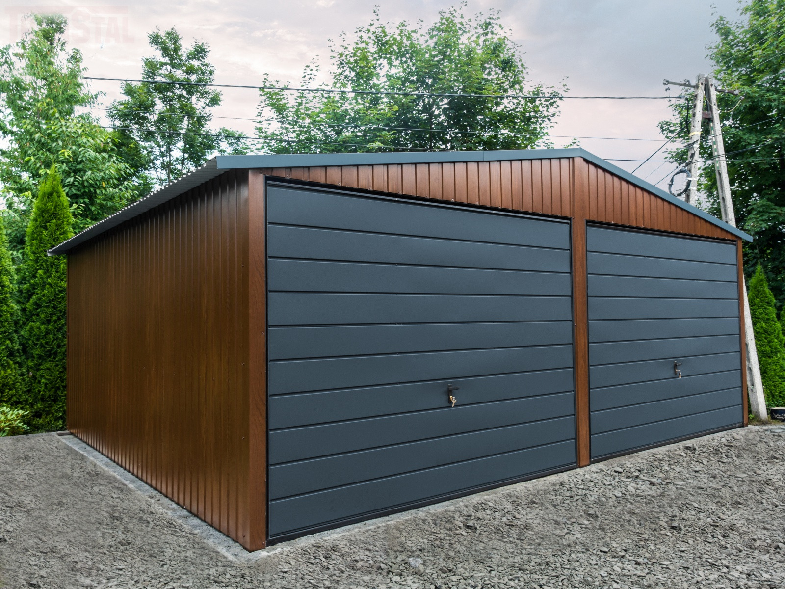 Dwustanowiskowy garaż blaszany z brązowymi ścianami i grafitowymi bramami segmentowymi, widoczny na tle zieleni.