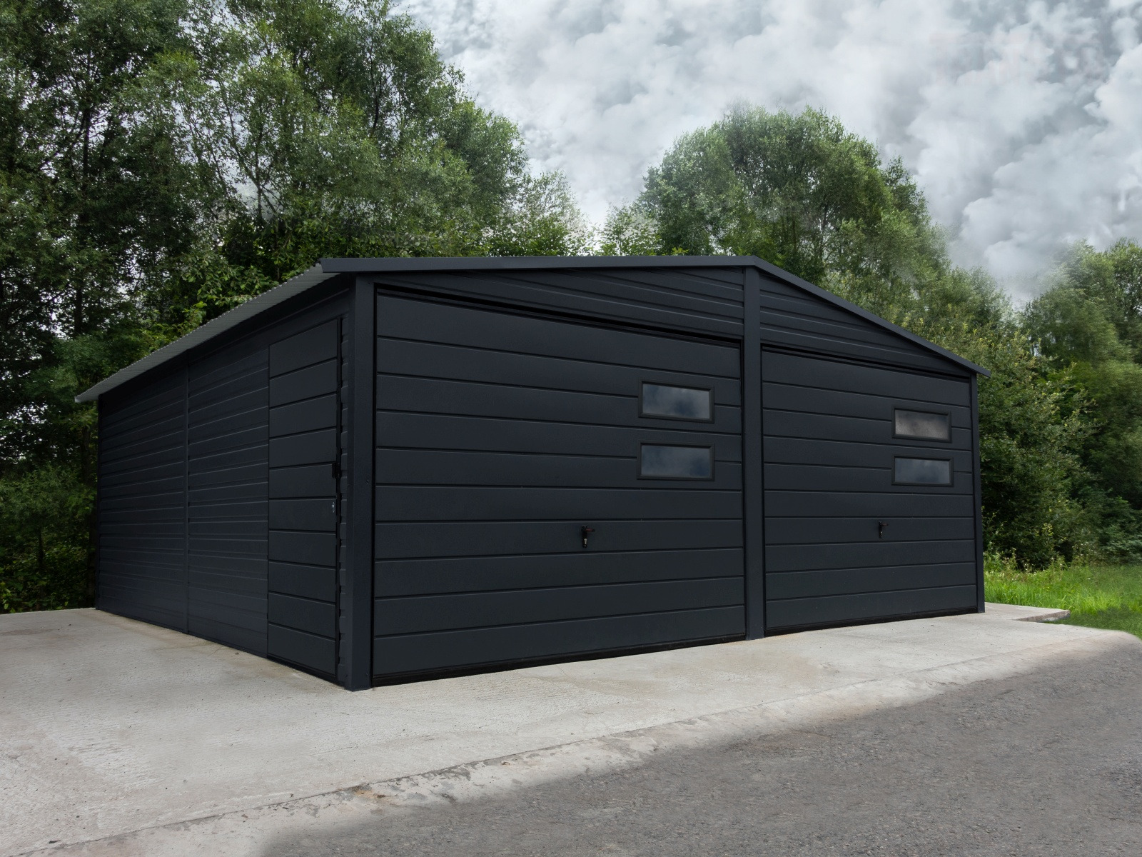 Dwustanowiskowy, ciemnoszary garaż blaszany z dwoma oknami na każdej bramie, usytuowany na betonowej wylewce, otoczony zielenią.