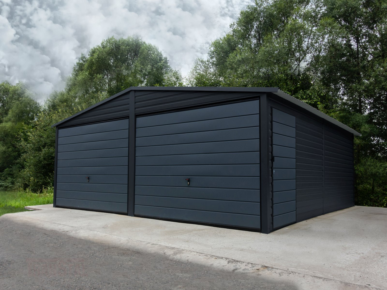Dwustanowiskowy, grafitowy garaż blaszany z poziomymi przetłoczeniami, umieszczony na betonowej wylewce, na tle zieleni i pochmurnego nieba.