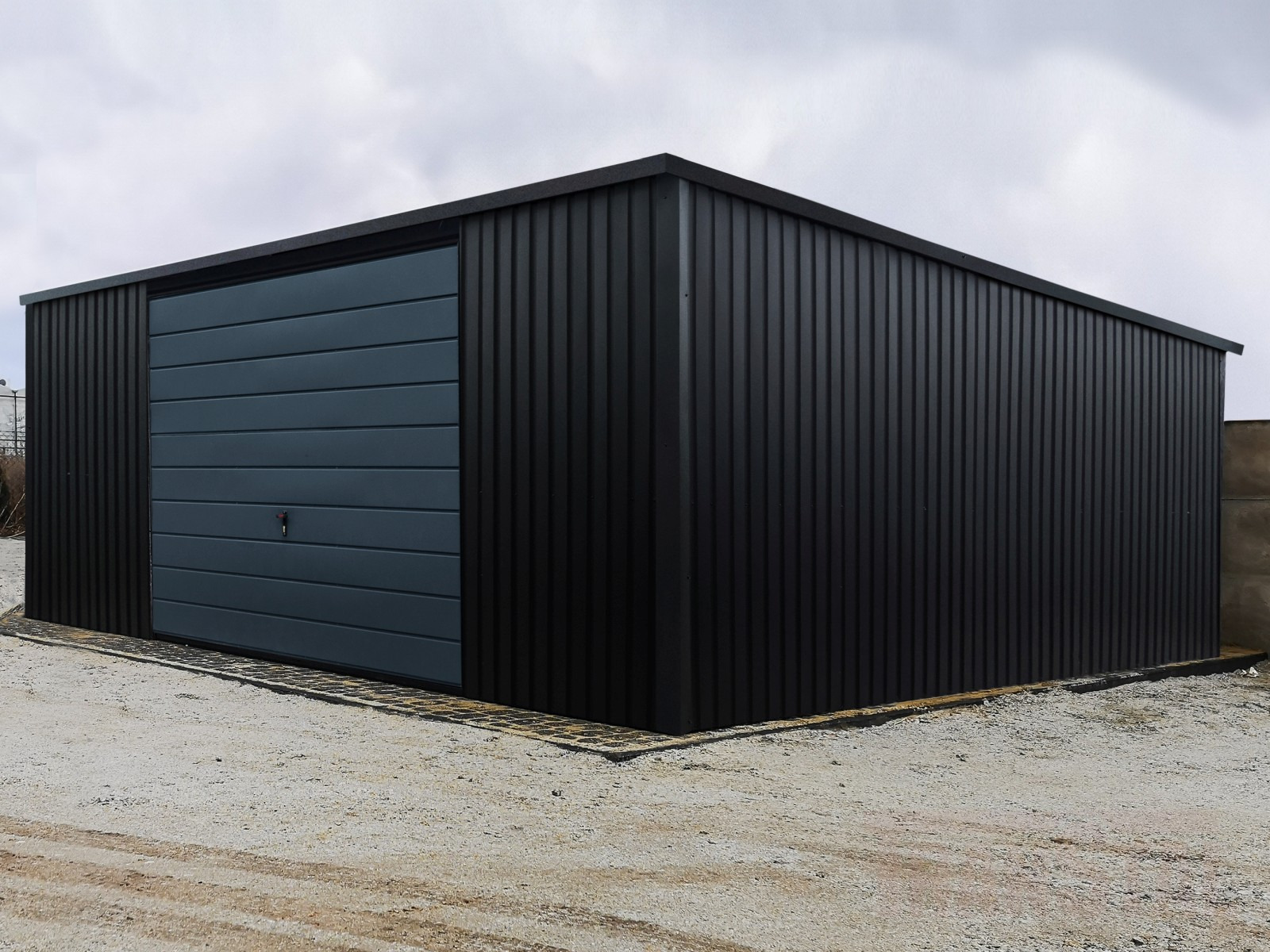 Nowoczesny, czarny garaż blaszany z szarą bramą segmentową, wykończony kostką brukową, na żwirowym podłożu, pod pochmurnym niebem.