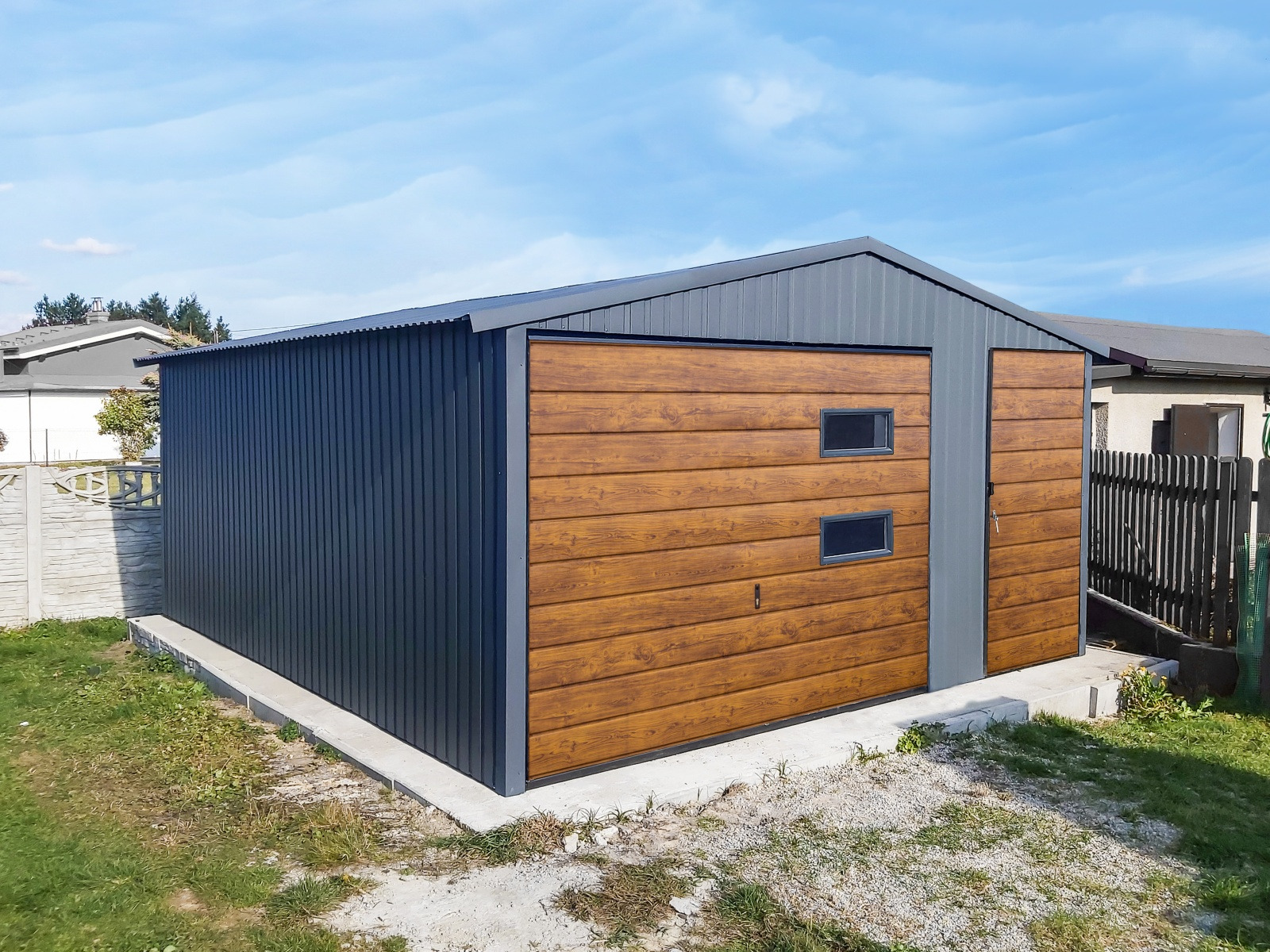 Nowoczesny garaż blaszany z bramą o fakturze drewna i dwoma małymi okienkami, ustawiony na betonowej wylewce, na tle trawnika i zabudowań.