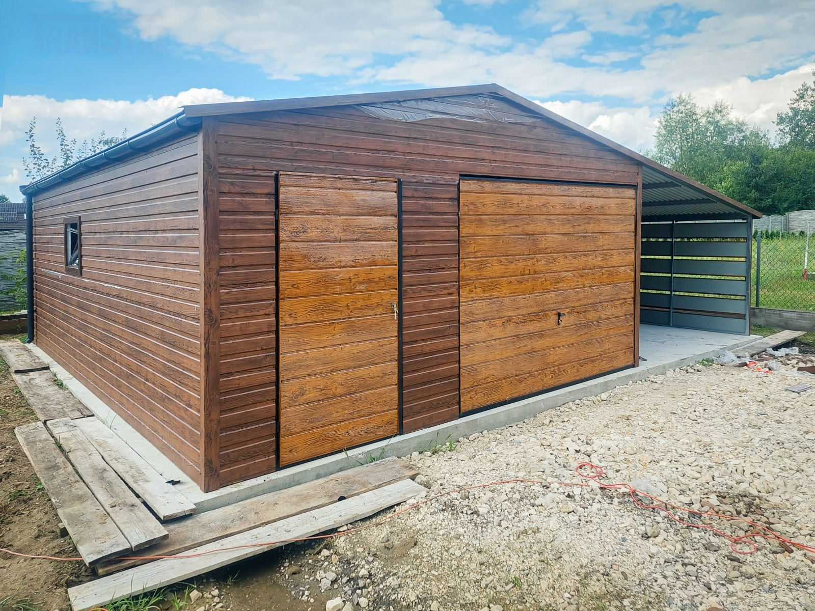 Brązowy garaż blaszany z imitacją drewna, z dwoma przesuwnymi drzwiami, małym oknem po lewej stronie i betonową wylewką, widoczny fragment zadaszenia z tyłu, na ziemi gruz i deski.