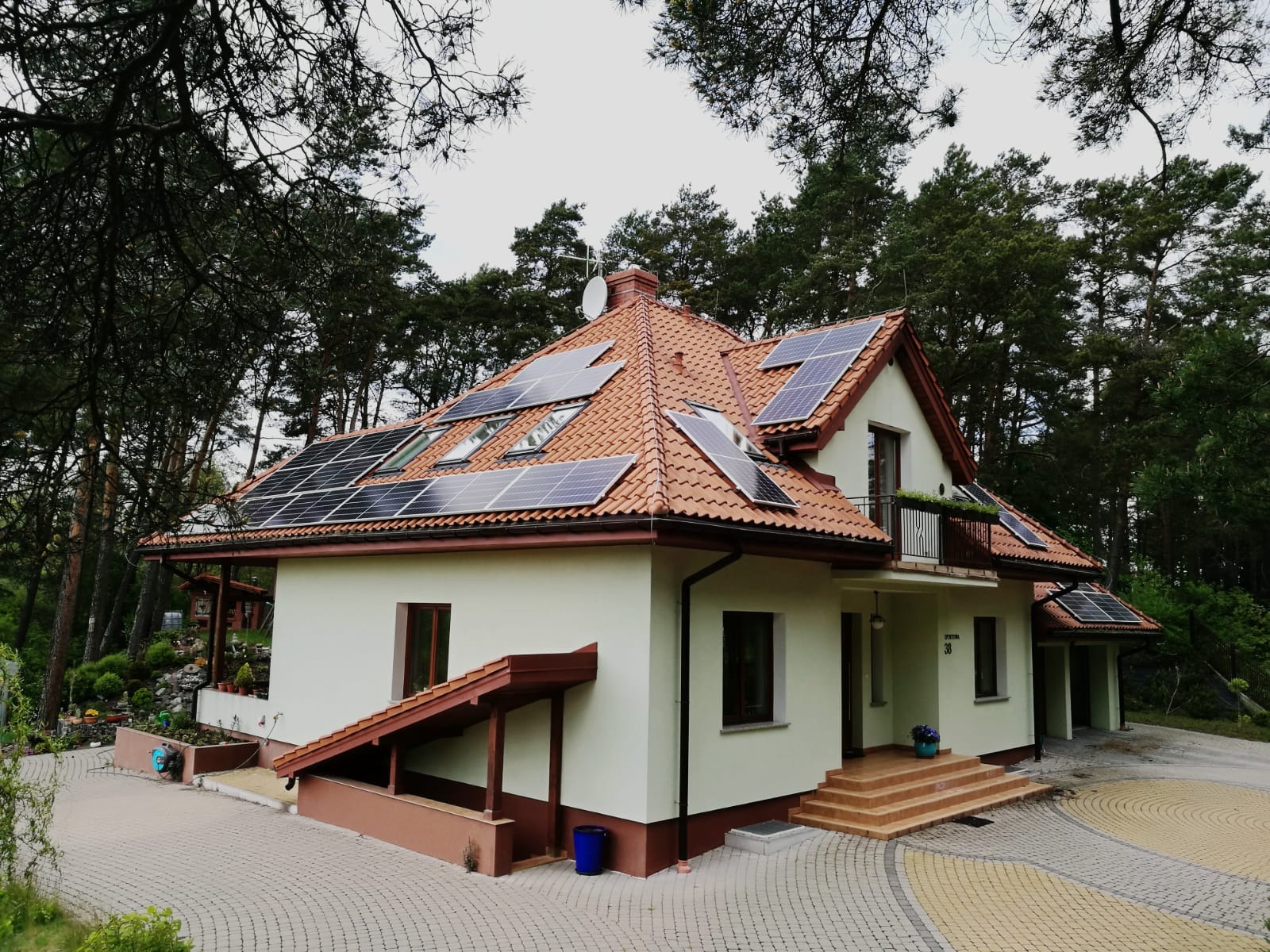 Dom jednorodzinny z panelami słonecznymi na dachu z czerwonej dachówki, widoczne okna dachowe, antena satelitarna, oraz drzewa w tle.