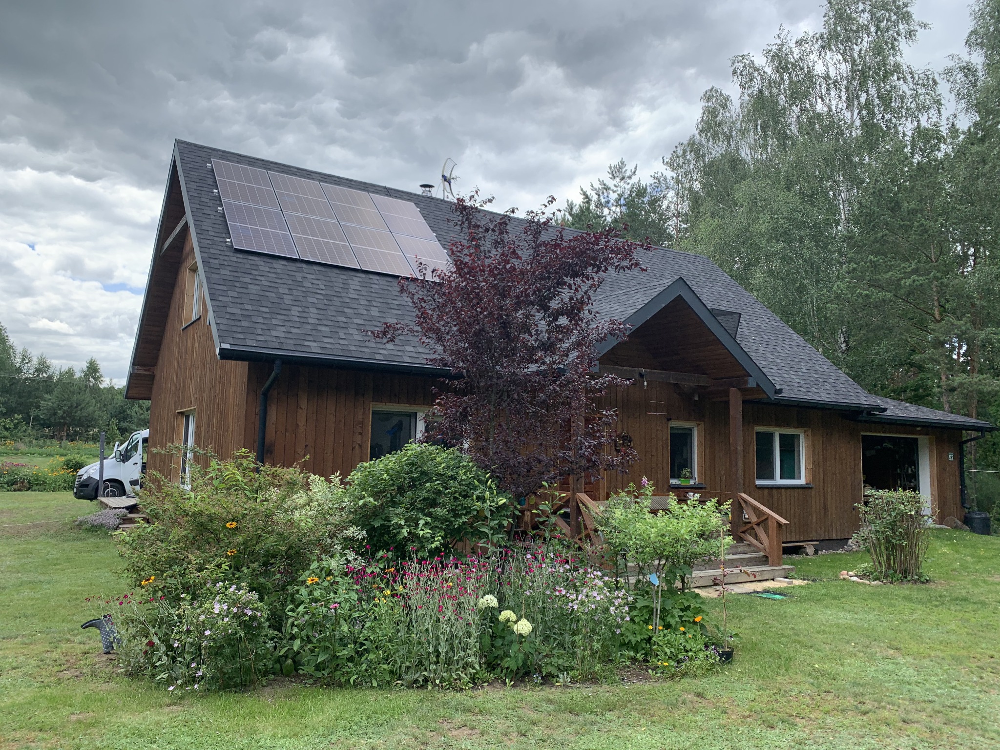 Brązowy dom jednorodzinny z ciemnoszarą dachówką i panelami słonecznymi na dachu, otoczony zielenią i drzewami, z widocznym białym vanem zaparkowanym z boku.