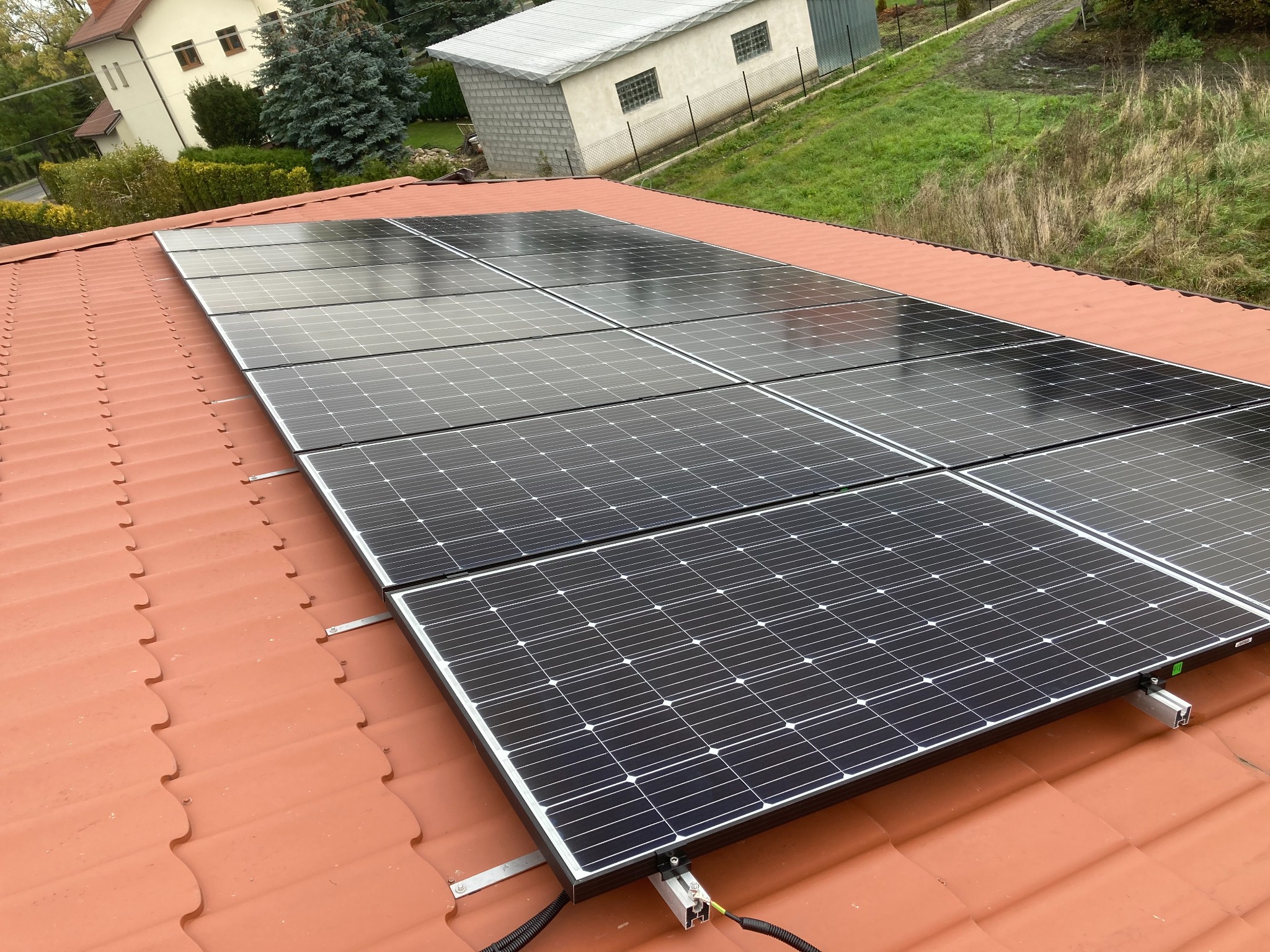 Panele słoneczne zamontowane na pomarańczowym dachu z dachówki ceramicznej, widoczne kable i elementy montażowe, w tle budynki i zieleń.