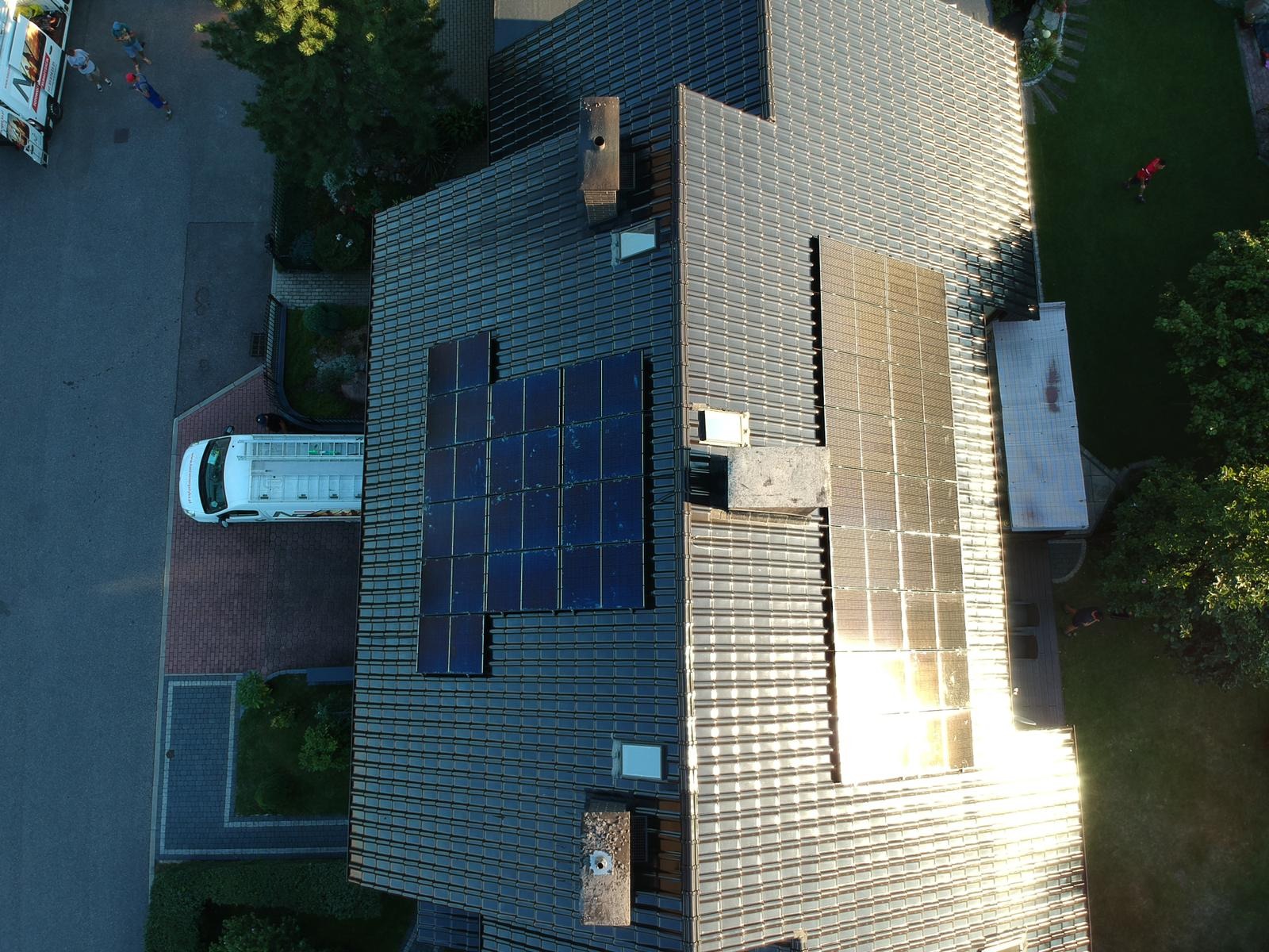 Widok z góry na dach domu z zamontowanymi panelami słonecznymi, obok zaparkowany biały bus serwisowy z drabiną na dachu, widoczny fragment chodnika i trawnika.