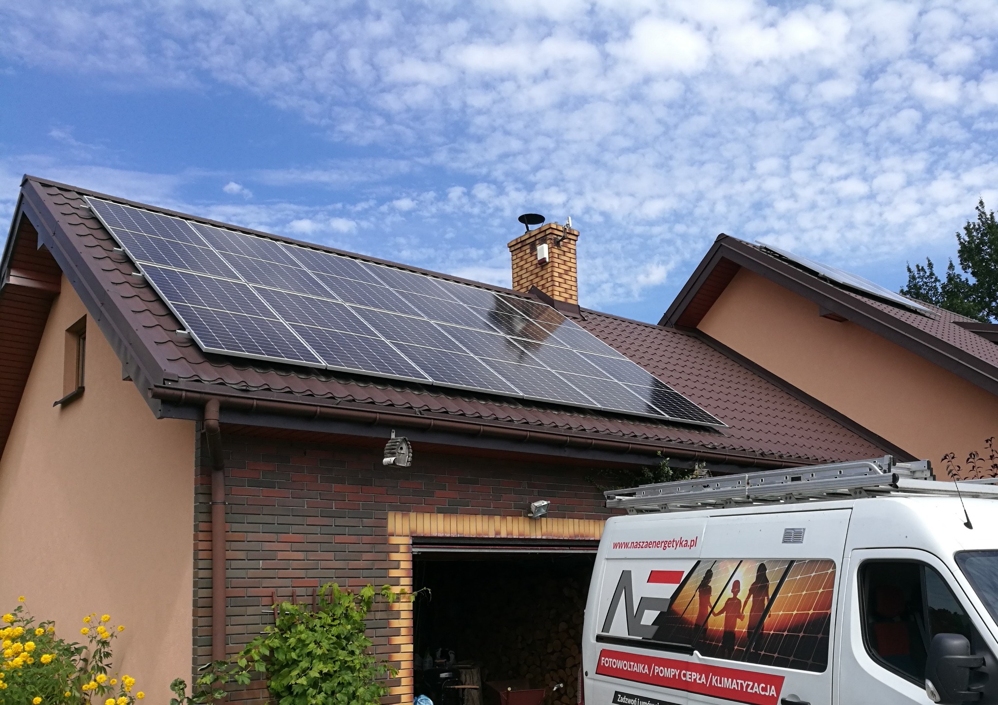 Panel fotowoltaiczny na brązowym dachu domu jednorodzinnego z ceglanym kominem, na tle błękitnego nieba z lekkimi chmurami. Biały samochód dostawczy z logotypem firmy instalacyjnej częściowo...