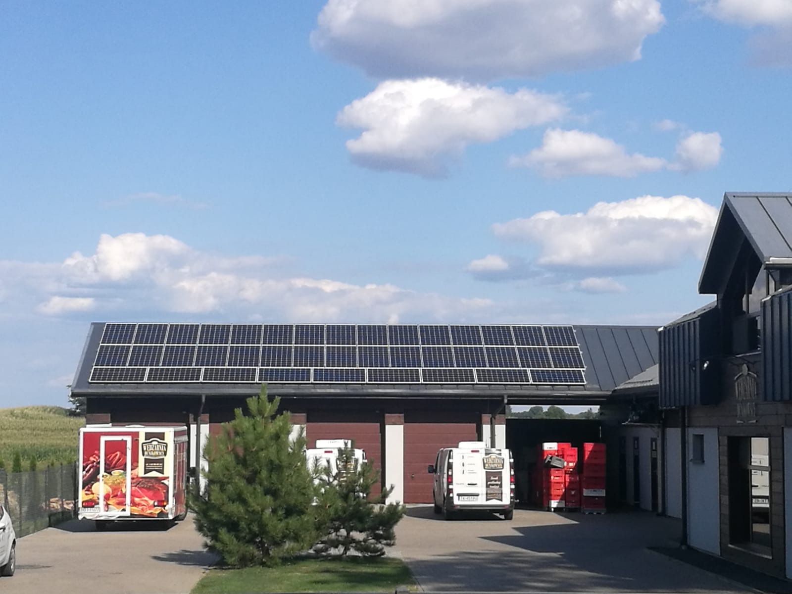 Panele słoneczne na dachu budynku gospodarczego, z zaparkowanymi samochodami dostawczymi i przyczepą chłodniczą w pobliżu.