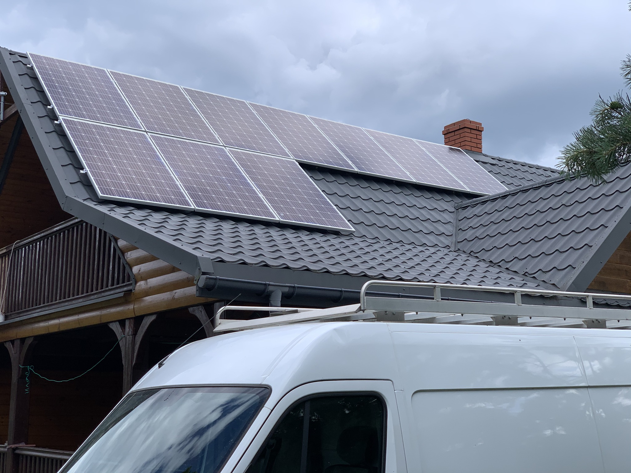 Panele fotowoltaiczne zamontowane na szarym dachu domu z bali, widoczny biały samochód dostawczy z bagażnikiem dachowym.