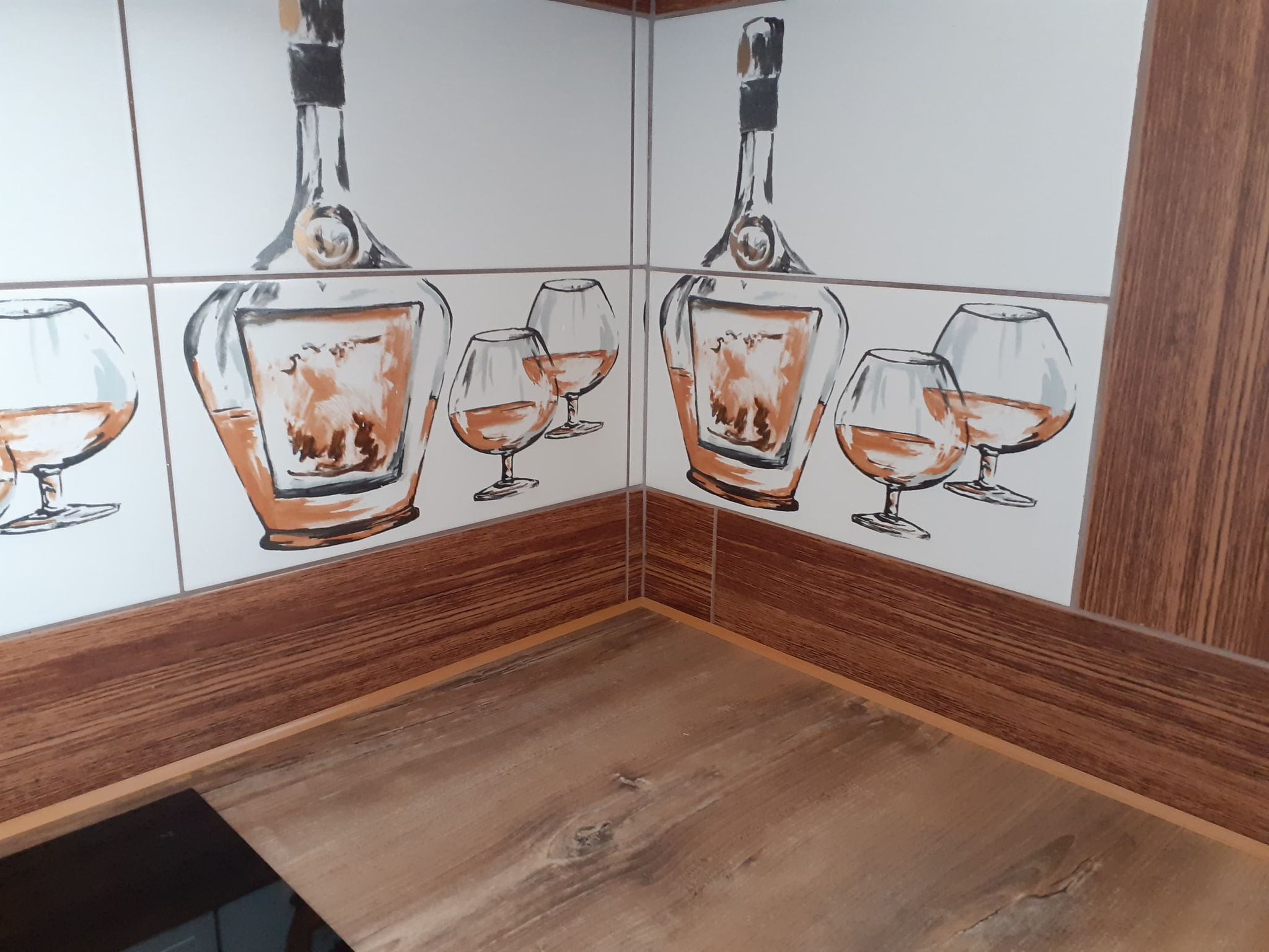 Wykończony narożnik kuchni z płytkami przedstawiającymi butelki i kieliszki, imitacja drewna na blacie i cokole.